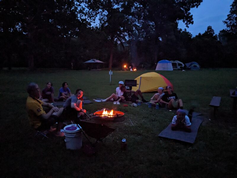In a grassy field at dusk, a group gathers around a campfire. Tents are pitched in the background, suggesting an overnight camping trip. The fire casts a warm glow on the people nearby, some sitting on the ground, others tending to the flames. The atmosphere appears relaxed and communal, with the focus on enjoying the evening outdoors.